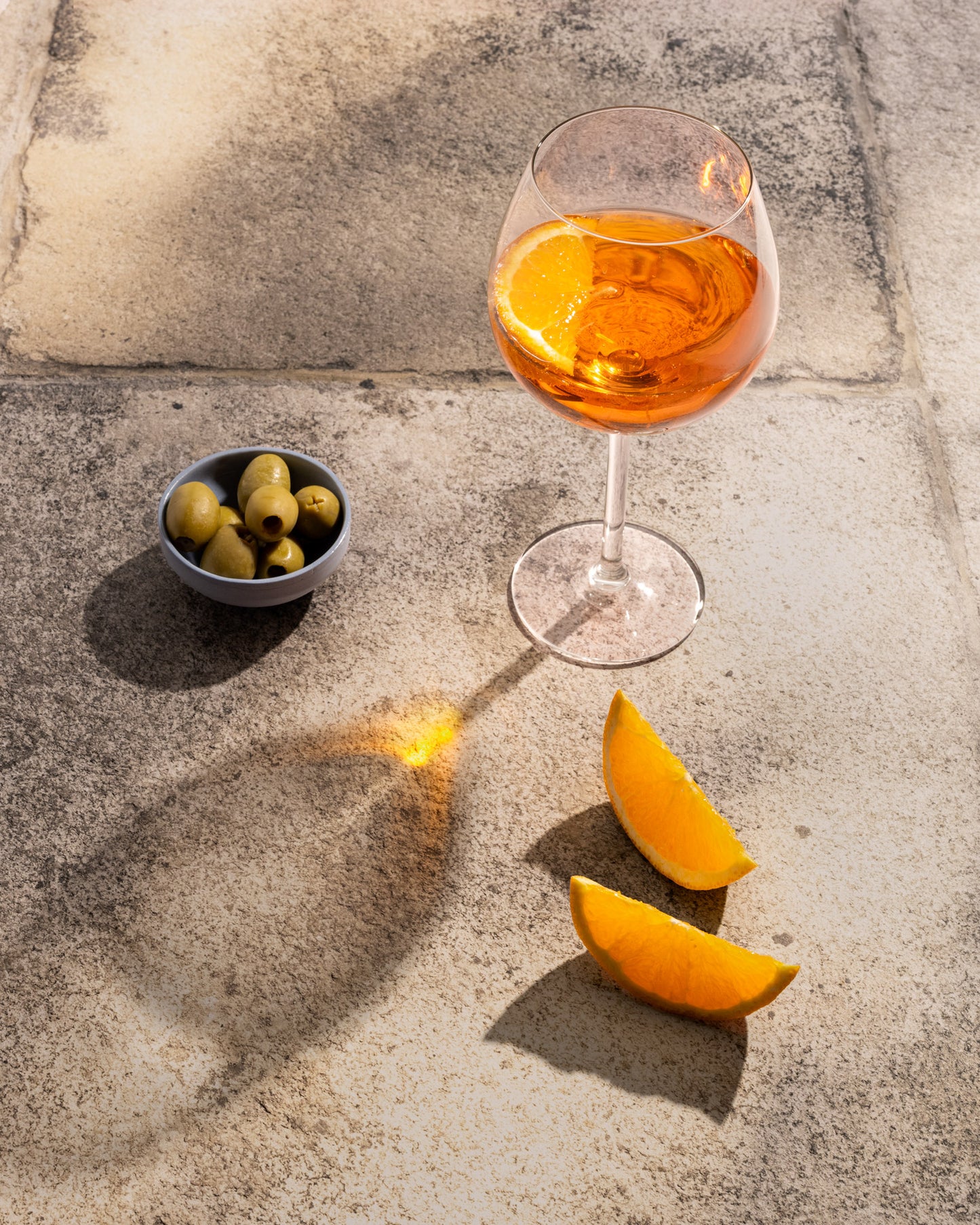 Wine glass with orange liquid on a stone surface with olives and orange slices.